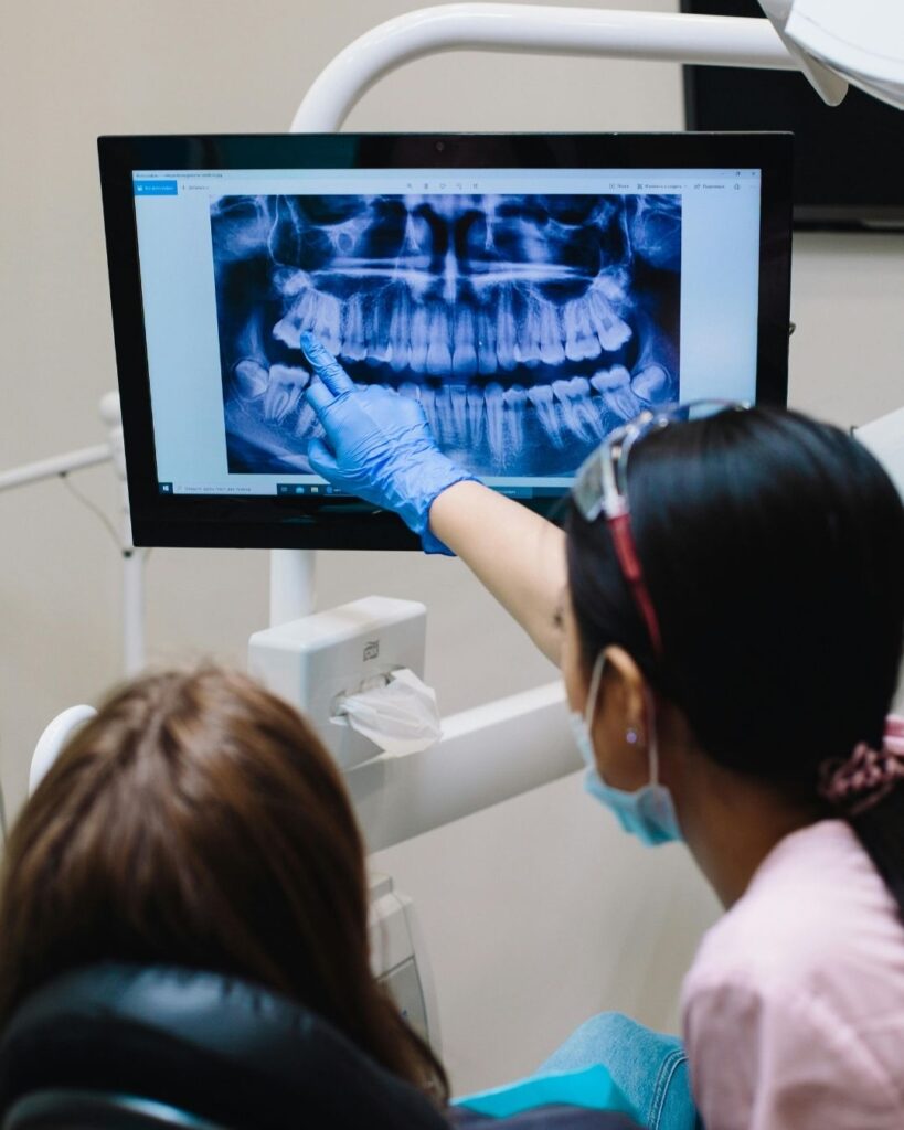 Female dentist explaining digital dental X-ray results to a patient while she lies comfortably in the dental chair.